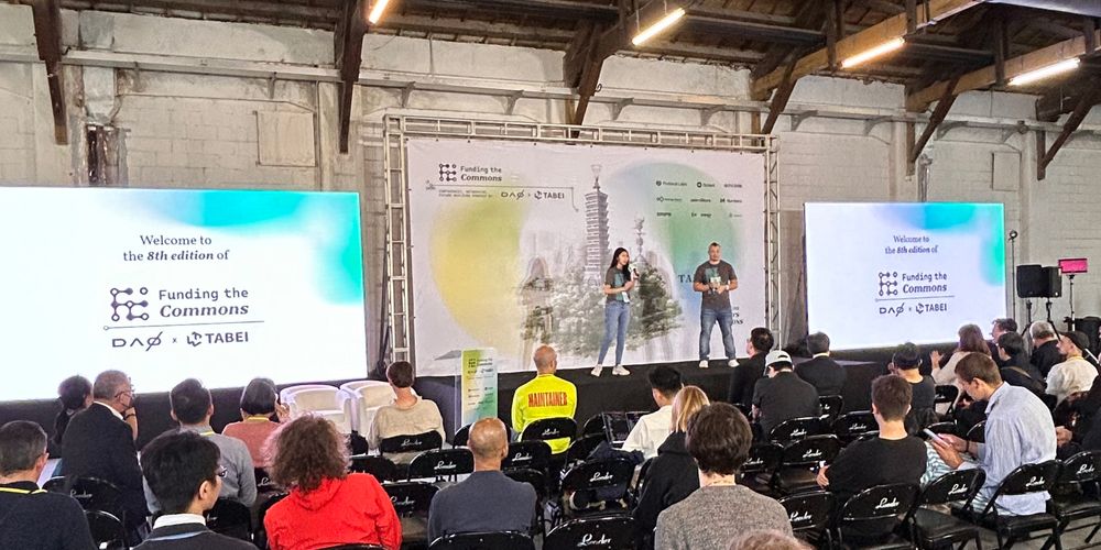 Two presenters stand on a stage in front of several screens in a warehouse in Taipei in front of a crowd 